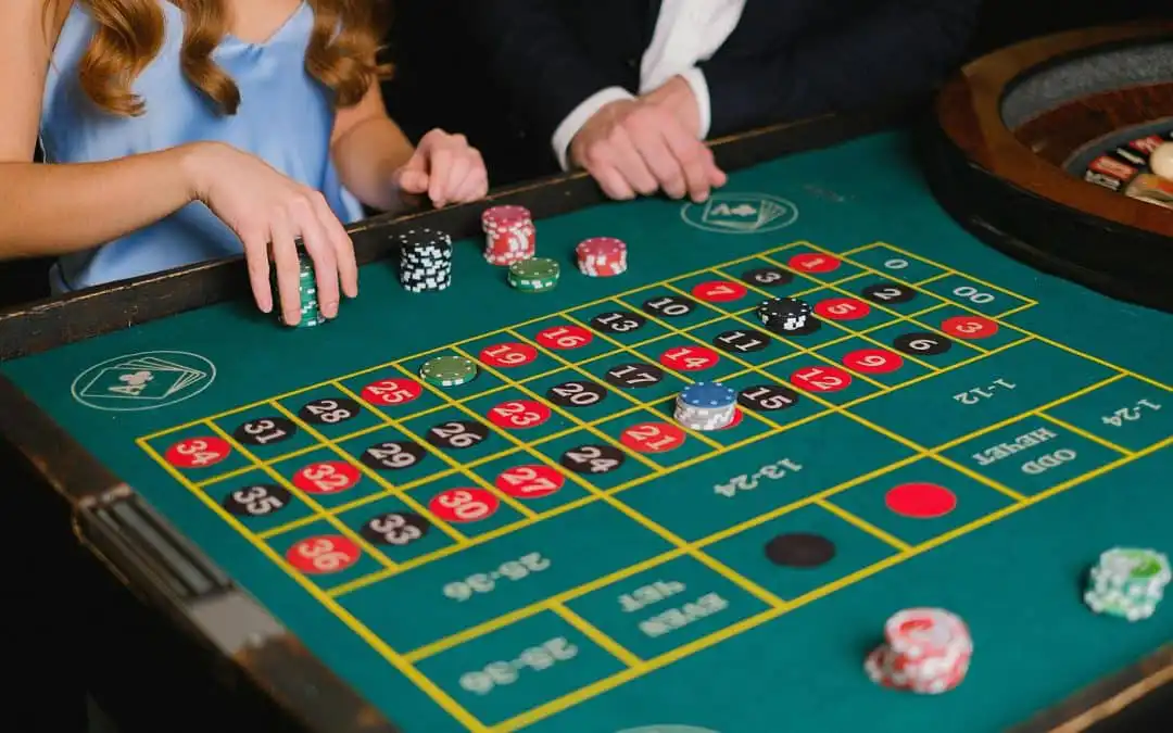 Two players place chips on a green roulette table with the numbered layout and betting areas.